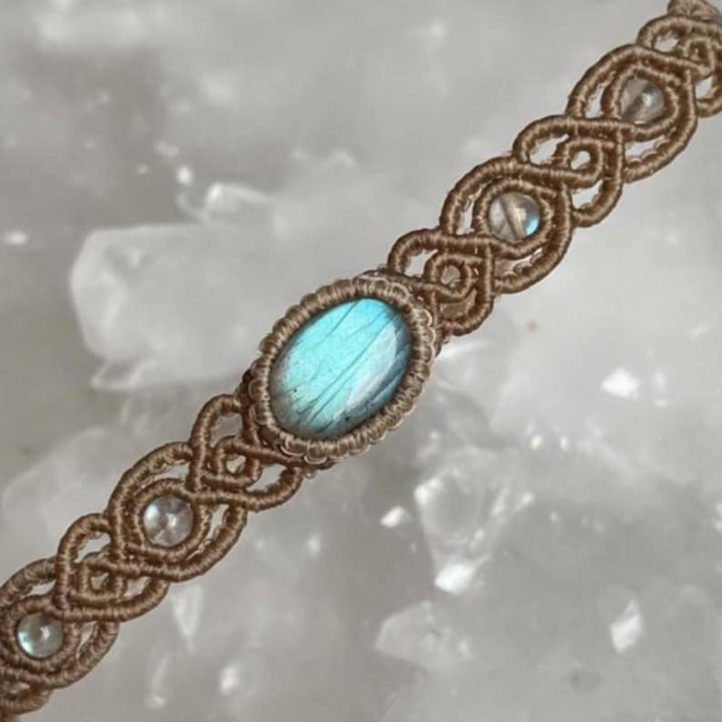 May include: A handmade macrame bracelet featuring a central oval labradorite stone with blue and green iridescence. The bracelet is woven with tan-colored cord and includes small, clear beads. The intricate knotwork creates a decorative pattern.