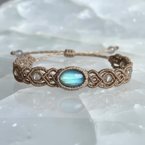 May include: A macrame bracelet featuring a central oval-shaped, iridescent blue gemstone. The bracelet is crafted with tan-colored cord, with clear beads interspersed throughout the design. The adjustable closure allows for a comfortable fit.