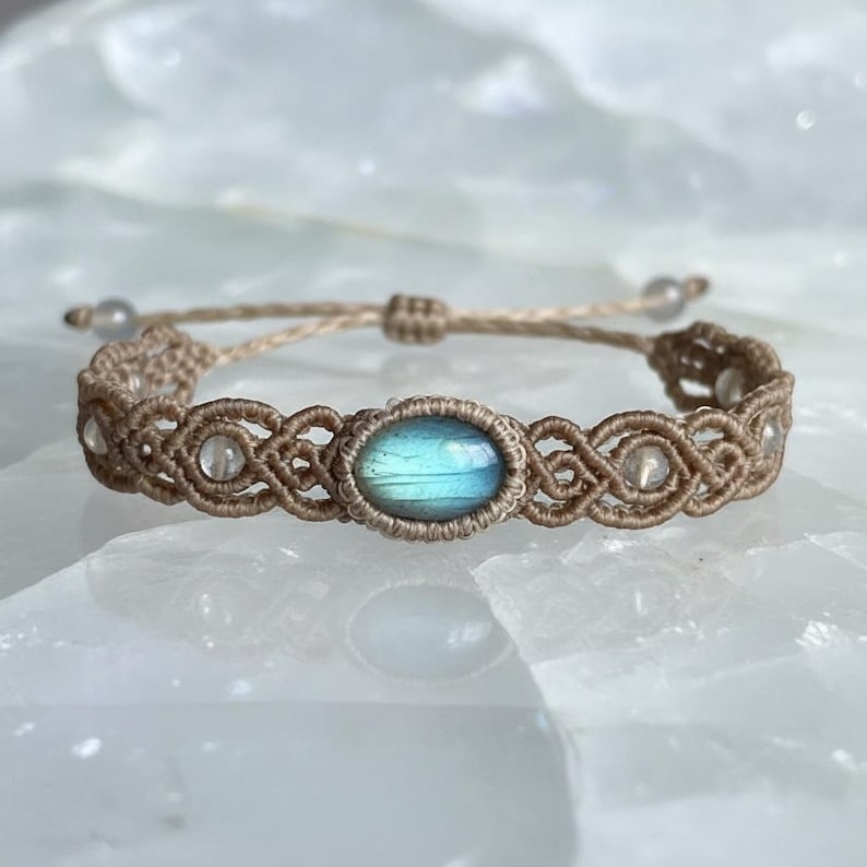 May include: A macrame bracelet featuring a central oval-shaped, iridescent blue gemstone. The bracelet is crafted with tan-colored cord, with clear beads interspersed throughout the design. The adjustable closure allows for a comfortable fit.