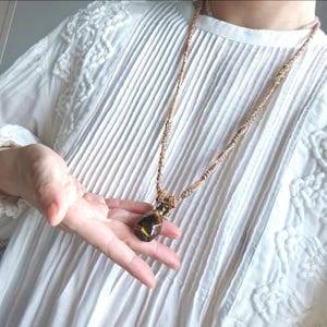 Puede incluir: Un collar artesanal con un colgante de piedra preciosa marrón y amarilla facetada. El colgante cuelga de un cordón trenzado con hilos beige, marrón y rojo. El collar se muestra sobre un fondo blanco texturizado.