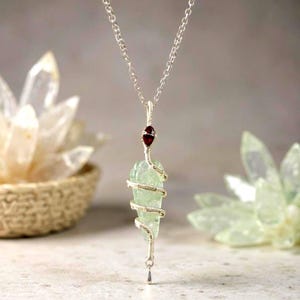 以下が含まれることがあります： 銀色のネックレス。淡い緑色の原石ペンダントが銀色のワイヤーで包まれています。小さな赤い宝石がクリスタルの上にセットされています。ネックレスは繊細な銀色のチェーンから吊り下げられています。背景にはクリスタルと織りのバスケットがあります。