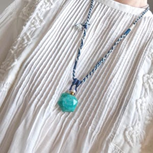Puede incluir: Collar con colgante de piedra turquesa y cordón tejido azul y blanco. El colgante tiene forma hexagonal facetada. El collar se muestra sobre una blusa blanca texturizada.