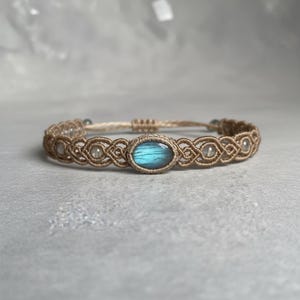 May include: A macrame bracelet featuring a central oval-shaped, blue-green gemstone. The bracelet is crafted with tan-colored cord, showcasing intricate knotting patterns. Small, clear beads are interspersed along the design, adding subtle highlights to the jewelry.