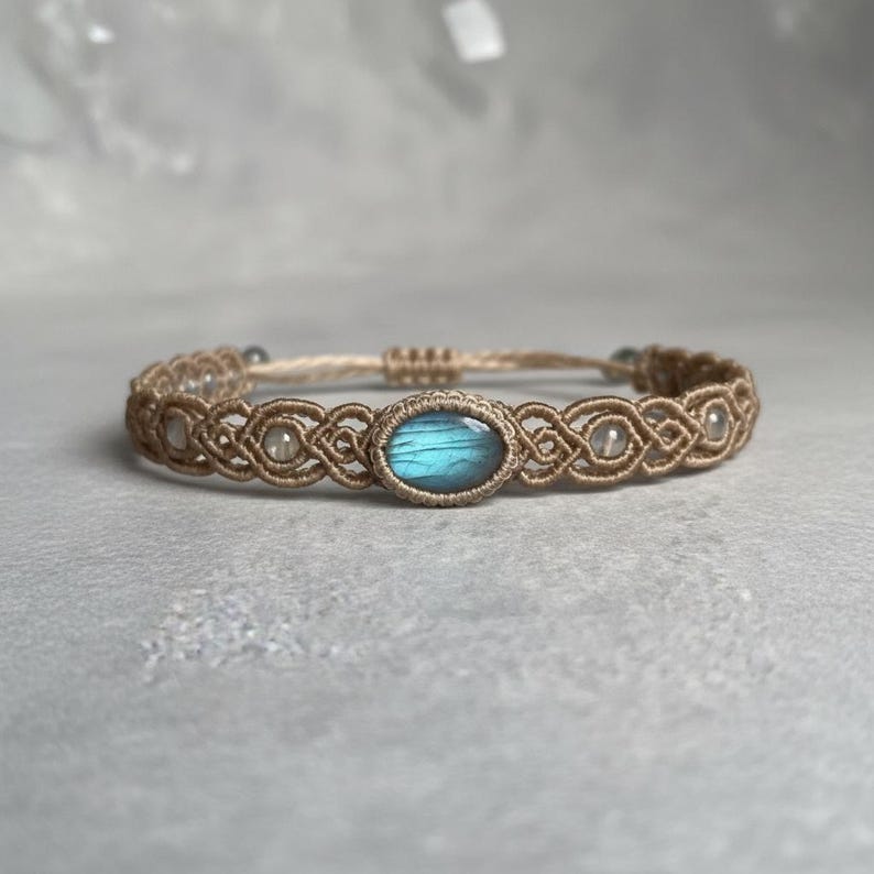 May include: A macrame bracelet featuring a central oval-shaped, blue-green gemstone. The bracelet is crafted with tan-colored cord, showcasing intricate knotting patterns. Small, clear beads are interspersed along the design, adding subtle highlights to the jewelry.