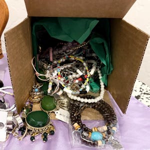 May include: A cardboard box filled with assorted jewelry including beaded bracelets, pearl strands, and rings. The box contains green tissue paper and various items such as a large green faceted stone pendant, dark brown beaded bracelets, and metal rings.