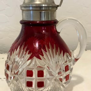 Può includere: Una caraffa in vetro tagliato rosso rubino e trasparente con un motivo geometrico a rombi e ventagli. Il contenitore presenta un manico in vetro trasparente e un coperchio in metallo argentato con una piccola leva sul bordo superiore.