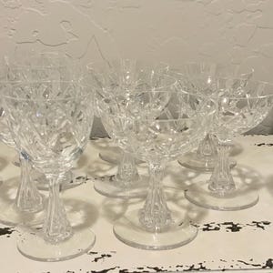 Può includere: Una collezione di bicchieri di cristallo trasparente, tra cui calici da vino e flûte da champagne, con intricati motivi a taglio diamante. I bicchieri sono disposti su una superficie bianca usurata, mettendo in risalto il loro design elegante.