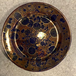 May include: A round, decorative plate with a metallic gold finish and a pattern of dark blue, irregular circles. The plate has a glossy surface and a slightly raised rim. The design gives the plate a unique, artistic appearance.