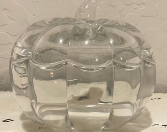 May include: A clear glass pumpkin-shaped container with a lid and stem. The container has a ribbed design and is sitting on a white surface. The pumpkin is a decorative item.