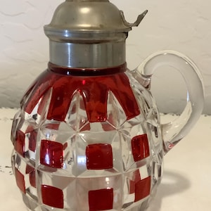 May include: A clear glass pitcher featuring a faceted diamond pattern with alternating red square accents. The pitcher has a rounded body, a clear glass handle, and a metallic silver-toned lid with a small finial and a hinged thumb lever.