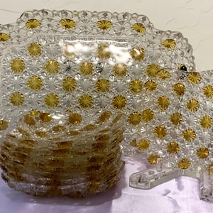 May include: A set of clear and amber glass dishes featuring a pressed starburst pattern. The collection includes one large rectangular platter, one square plate on a clear stand, and a stack of smaller square plates, all with gold-toned centers.