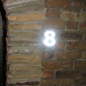 Könnte beinhalten: Eine leuchtend weiße Zahl Acht ist an einer Backsteinmauer angebracht. Die Mauer besteht aus braunen Ziegeln und Steinen. Die Zahl leuchtet hell und bildet einen Kontrast zum dunkleren Hintergrund.