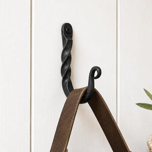 May include: A black, wrought iron hook with a twisted design is mounted on a white wooden wall. A brown leather strap of a bag hangs from the hook. A woven basket and greenery are in the background.