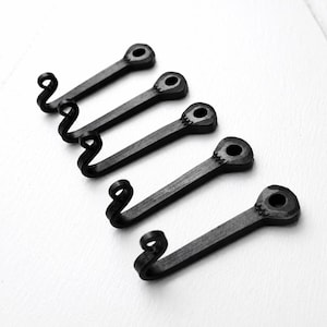 Könnte beinhalten: Fünf schwarze Metallhaken, diagonal angeordnet. Jeder Haken hat einen rechteckigen Körper mit einem Loch und einem gebogenen Hakenende. Diese Haken sind wahrscheinlich zum Aufhängen von Gegenständen gedacht.