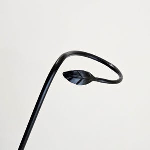 Op de afbeelding: Een zwarte metalen plantenstaak met een bladvormgeving. De staak heeft een gebogen bovenkant en een klein, gedetailleerd blad aan het einde van de bocht. Het blad heeft zichtbare nerven en is effen zwart.