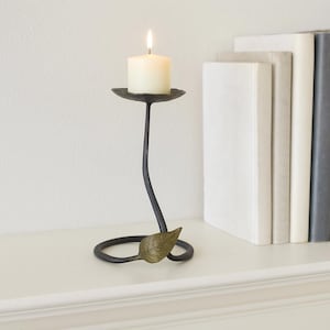 Puede incluir: Un candelabro de metal negro con una vela blanca encendida. El soporte tiene un tallo curvo y un adorno en forma de hoja. Una pila de libros está en el fondo. El candelabro mide aproximadamente 25 cm de alto.
