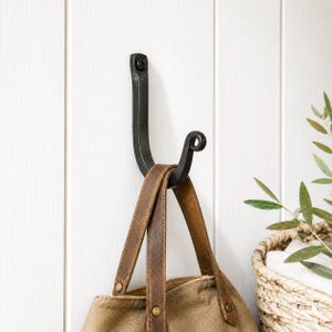 May include: A black, wrought iron hook mounted on a white, vertical-paneled wall. The hook is holding a canvas tote bag with brown leather straps. A decorative scroll detail is at the end of the hook.
