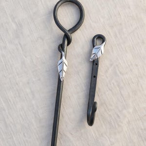 May include: Two black wrought iron fireplace tools. One tool is a poker with a loop handle and a silver leaf accent. The other is a hook with a silver leaf accent. Both tools are on a neutral background.
