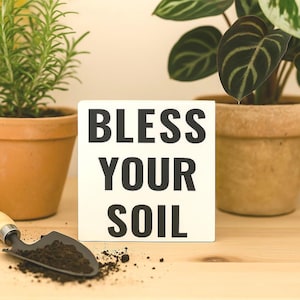 May include: A square, off-white sign with the words "BLESS YOUR SOIL" in bold, black letters. A small gardening trowel with soil sits in front of the sign. Potted plants are in the background.