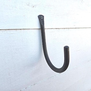 Könnte beinhalten: Ein dunkelgrauer Schmiedeeisenhaken, montiert auf einer weißen Holzoberfläche. Der Haken ist gebogen mit einem abgerundeten Ende und wird mit einer einzigen Schraube befestigt. Das Design ist schlicht und rustikal.