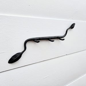May include: Black metal wall hook with a leaf and branch design. The hook has three curved hooks and a leaf-shaped end. The hook is mounted on a white wooden surface.