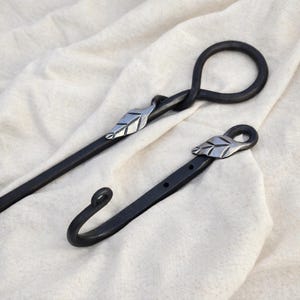 May include: Two black metal fireplace tools. One tool has a looped handle and a leaf accent. The other tool is a hook with a leaf accent. Both tools are on a white fabric background.