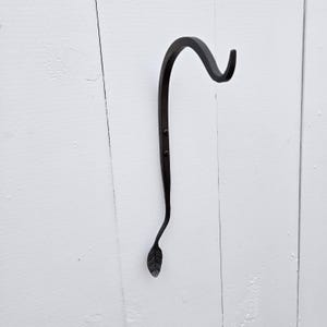 Op de afbeelding: Een zwarte, handgesmede metalen haak met een bladontwerp aan de onderkant. De haak buigt omhoog en heeft drie klinknagels langs de steel. De haak is gemonteerd op een wit houten oppervlak.
