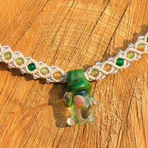 May include: A handmade macrame necklace with a green glass pendant. The necklace features a cream-colored woven cord with green and yellow beads. The pendant is a small, green glass figure with internal details, resting on a wooden surface.