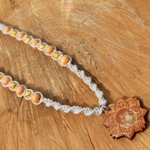 Könnte beinhalten: Eine handgefertigte Halskette mit einem hellgrauen Makramee-Band, Holzperlen und goldfarbenen Akzenten. Die Halskette hat einen Holzanhänger mit einem komplizierten Design und einem zentralen Stein. Die Halskette wird auf einer Holzoberfläche präsentiert.