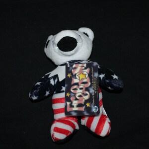 May include: A white plush bear wearing a red, white and blue American flag costume. The bear is holding a tag that says "For Your Home" and "Grateful Dead Bear for collectors by Pizza Bite".