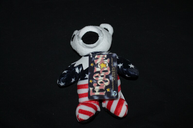 May include: A white plush bear wearing a red, white and blue American flag costume. The bear is holding a tag that says "For Your Home" and "Grateful Dead Bear for collectors by Pizza Bite".