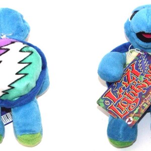 Puede incluir: Tortuga de peluche azul con una concha verde, blanca y morada con el logotipo Steal Your Face de los Grateful Dead. La tortuga lleva una camisa a rayas amarilla y verde y tiene una etiqueta que dice "Lazy Lightning".