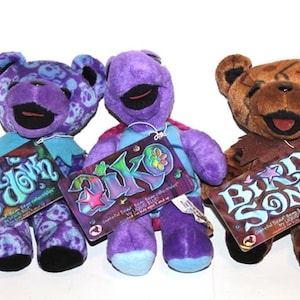 May include: Three purple and brown plush teddy bears with different designs. The bears are wearing colourful clothing and have tags that say "Shake & Down", "Diko", and "Bird Song".