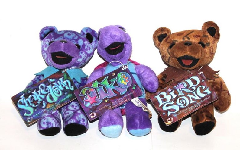 May include: Three purple and brown plush teddy bears with different designs. The bears are wearing colourful clothing and have tags that say "Shake & Down", "Diko", and "Bird Song".