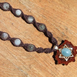 Puede incluir: Un collar de macramé de color marrón con un colgante en forma de estrella que presenta una piedra preciosa azul. El collar está adornado con varias piedras de color azul claro y gris.