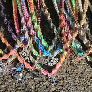 May include: A collection of colorful friendship bracelets with various silver charms, including a crescent moon, a dragonfly, a peace sign, a lotus flower, and an Om symbol.
