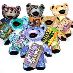 May include: A collection of seven colorful plush teddy bears with different patterns and designs. Each bear has a tag with a different name, including "Home", "Aloha", "Birthday Show", "Trixie", "Grateful", "Wander", and "Kona".