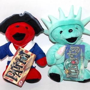 Puede incluir: Dos osos de peluche vestidos como figuras patri&oacute;ticas americanas. El oso rojo lleva un traje de marinero azul y blanco y un sombrero de pirata negro. El oso verde lleva un disfraz verde de la Estatua de la Libertad. Ambos osos tienen etiquetas que dicen "Patriot" y "Liberty" con una estrella y un esquema de color rojo, blanco y azul.