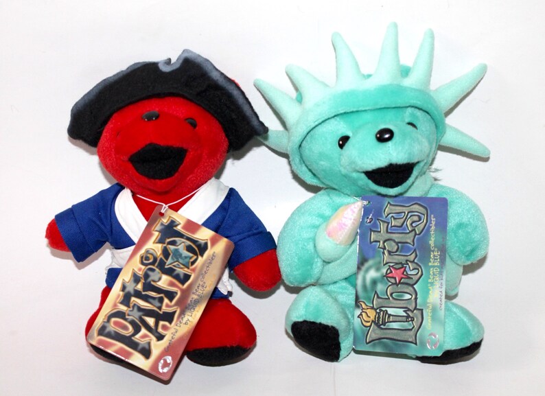 Puede incluir: Dos osos de peluche vestidos como figuras patri&oacute;ticas americanas. El oso rojo lleva un traje de marinero azul y blanco y un sombrero de pirata negro. El oso verde lleva un disfraz verde de la Estatua de la Libertad. Ambos osos tienen etiquetas que dicen "Patriot" y "Liberty" con una estrella y un esquema de color rojo, blanco y azul.