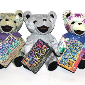 May include: Three plush teddy bears with different colourful patterns. The bears are wearing purple, grey, and blue collars. Each bear has a tag with a different design and text. The tags read "Eyes of the World", "Peace Moon", and "Master Peace".