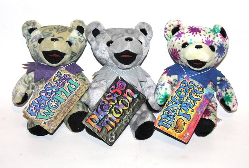 May include: Three plush teddy bears with different colourful patterns. The bears are wearing purple, grey, and blue collars. Each bear has a tag with a different design and text. The tags read "Eyes of the World", "Peace Moon", and "Master Peace".