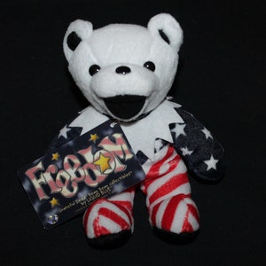 May include: A white plush bear wearing a red, white and blue American flag outfit. The bear is holding a tag that says "Freedom" with a star on it. The tag also says "Grateful Dead Bear from collectors by Liquid Blue".