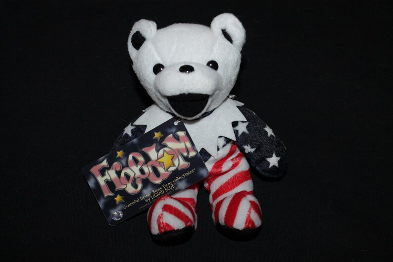 May include: A white plush bear wearing a red, white and blue American flag outfit. The bear is holding a tag that says "Freedom" with a star on it. The tag also says "Grateful Dead Bear from collectors by Liquid Blue".
