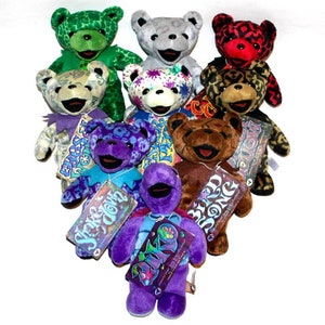 May include: A collection of ten colourful plush teddy bears with different patterns and designs. The bears are arranged in a heart shape with their tags visible. The tags feature the names of the bears, such as "Shakedown" and "Bird Song".