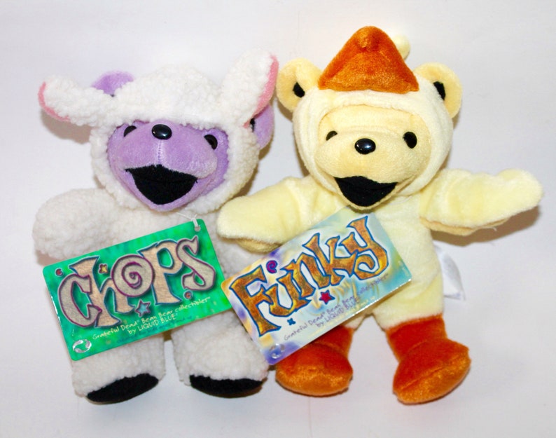 Puede incluir: Dos osos de peluche con disfraces. Un oso lleva un disfraz de oveja blanco con la cara morada y la etiqueta "CHOPS Grateful Dead Bear by Liquid Blue" adjunta. El otro oso lleva un disfraz de pollo amarillo con el pico naranja y la etiqueta "FINKY Grateful Dead Bear by Liquid Blue" adjunta.