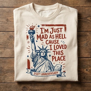 May include: A cream-colored t-shirt featuring a graphic of the Statue of Liberty holding a torch and a tablet. The text reads: I'm Just Mad As Hell Cause I Loved This Place, and 250th Anniversary. The design uses red and blue ink colors.