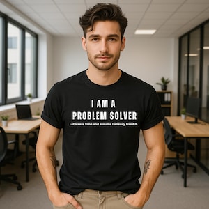 May include: Black t-shirt with the white text "I AM A PROBLEM SOLVER" and the phrase "Let's save time and assume I already fixed it." The t-shirt is worn by a person in an office environment.