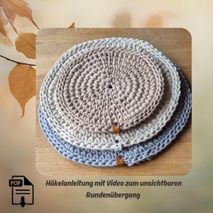 May include: Three crocheted circular placemats in beige, off-white, and light blue, stacked on top of each other. Each mat has a leather tag. The text "Häkelanleitung mit Video zum unsichtbaren Rundenübergang" is visible.