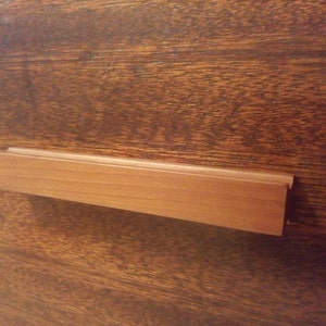 May include: A light brown wooden drawer pull with a rectangular shape and a grooved design. The pull is mounted on a dark brown wooden surface, showcasing the natural wood grain.