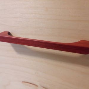 Pode incluir: Um puxador de arm&aacute;rio de madeira castanho-avermelhado com uma pega curva. O puxador &eacute; montado numa superf&iacute;cie de madeira clara. A pega tem aproximadamente 25 cm de comprimento.